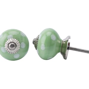 May include: Two green ceramic cabinet knobs with white polka dots. Each knob has a silver-coloured metal base and a threaded screw for installation. The knobs are designed for furniture and home decor.