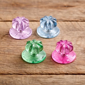 May include: Four colourful glass cabinet knobs in shades of purple, blue, green, and pink. Each knob features a flower-shaped top and a round base, set against a wooden background.