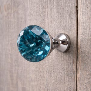 Op de afbeelding: Een ronde meubelknop met een gefacetteerde, briljant geslepen blauwe glazen bovenkant op een gepolijste chromen metalen voet. De knop heeft een cirkelvormig, meervoudig gefacetteerd oppervlak dat licht reflecteert, bevestigd aan een zilverkleurige steel.