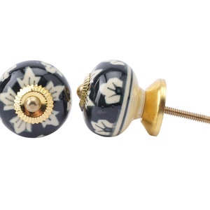 Puede incluir: Dos tiradores decorativos para armarios. Los tiradores son redondos con fondo negro y un estampado floral blanco. Tienen una base y un tornillo dorados. Los tiradores probablemente son de cerámica o porcelana.