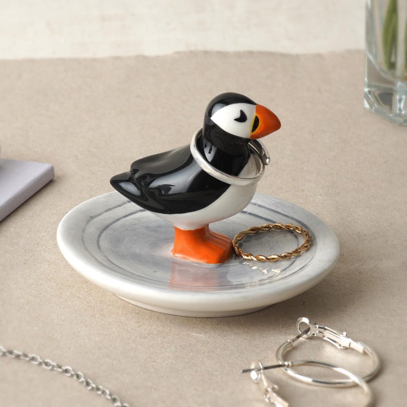 May include: A ceramic ring holder shaped like a puffin bird, with black, white, and orange coloring. The bird stands on a small, round, gray dish. A silver ring is around the bird's neck, and a gold ring rests on the dish.