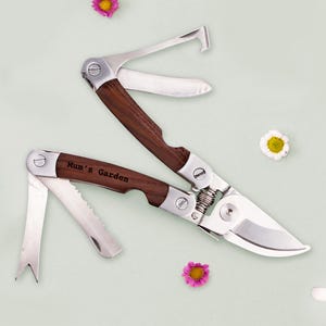 May include: A multi-tool gardening shear with a wooden handle and stainless steel blades. The text "Mum's Garden" is engraved on the handle. The tool is open, displaying various blades and a hook. The background is a light green with small flowers.