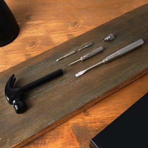 Puede incluir: Un martillo negro y un juego de destornilladores y puntas plateadas están dispuestos sobre una superficie de madera. Las herramientas están diseñadas para diversas tareas, mostrando un conjunto compacto y versátil para reparaciones y proyectos.