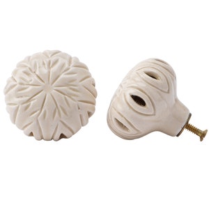 May include: Two white ceramic cabinet knobs with intricate carved designs. One knob is round with a starburst pattern, the other is a rounded oval with a floral design and a brass screw.