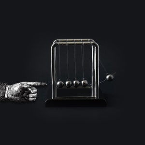 Op de afbeelding: Een Newton's cradle met vijf gepolijste stalen ballen die aan een chromen frame zijn opgehangen. Eén bal wordt teruggetrokken en losgelaten, wat de overdracht van momentum aantoont. Een geïllustreerde hand wijst naar de wieg tegen een donkere achtergrond.