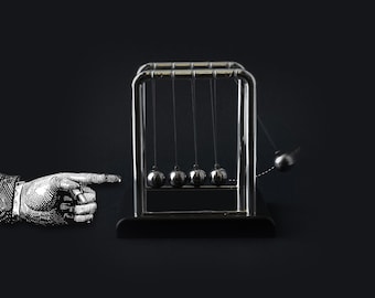 May include: A Newton's cradle with five polished steel balls suspended from a chrome frame. One ball is pulled back and released, demonstrating the transfer of momentum. An illustrated hand points towards the cradle against a dark background.