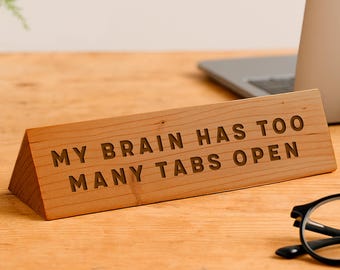May include: A wooden desk sign with the engraved text "MY BRAIN HAS TOO MANY TABS OPEN." The sign is a light brown color and is angled, resting on a wooden desk. A laptop and glasses are in the background.