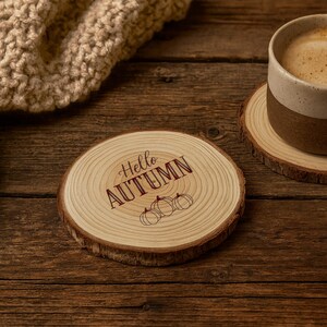 May include: A wooden coaster with the text "Hello AUTUMN" and three pumpkin illustrations. The coaster is made from a cross-section of a tree branch, with a natural bark edge. A mug of coffee sits on a similar coaster.