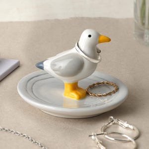May include: A ceramic ring holder shaped like a seagull, with a white body, gray wings, and yellow feet and beak. The seagull stands on a round, marbled dish. A silver ring is on the seagull's neck, and a gold ring rests on the dish.