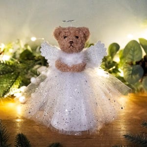 May include: A brown teddy bear angel tree topper with white wings and a white tulle skirt with silver glitter. The bear is wearing a halo.