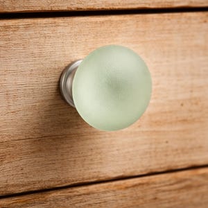 Könnte beinhalten: Ein kugelförmiger Möbelknopf mit einer mattierten, blass minzgrünen Glasoberfläche. Der Knopf hat eine glatte, abgerundete Form und ist an einem runden Sockel aus gebürstetem silberfarbenem Metall befestigt, der direkt an der Holzfront sitzt.