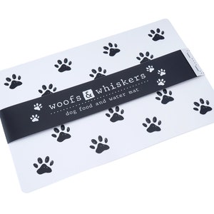 Puede incluir: Alfombra blanca para comida y agua para perros con huellas de patas negras y el texto "Woofs & Whiskers dog food and water mat".