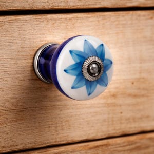 May include: A round ceramic drawer knob featuring a white surface with a painted blue floral pattern. The center of the flower is accented with a small, circular, ridged silver metal detail. The base of the knob is a smooth, dark blue ceramic.