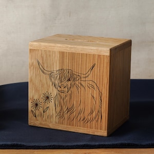 Puede incluir: Una caja de madera con un diseño tallado de una vaca de las Tierras Altas y dientes de león. La caja tiene una ranura en la parte superior para monedas o billetes.