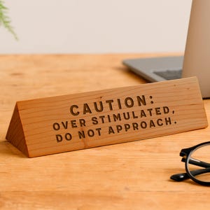 Op de afbeelding: Een houten bureau-bord met de tekst "CAUTION: OVER STIMULATED, DO NOT APPROACH." Het bord is gemaakt van lichtgekleurd hout en heeft een driehoekige vorm. De tekst is donker gegraveerd. Het bord staat op een houten bureau.