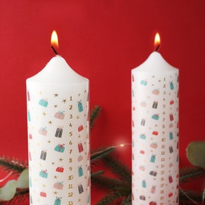 Puede incluir: Dos velas de adviento blancas con un patrón dorado y blanco de estrellas y cajas de regalo. Las velas están numeradas del 1 al 13 y del 1 al 19.