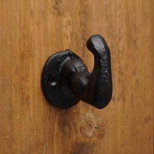 May include: A black, cast iron hook mounted on a wooden surface. The hook has a curved design and a textured finish. The mounting plate is round and also black.
