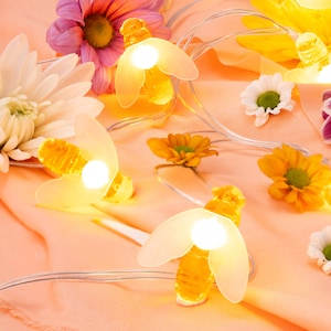 May include: A string of fairy lights with bee-shaped bulbs, strung on a peach-coloured fabric background. The lights are lit and glowing, and there are several white, yellow, and pink flowers scattered around the lights.