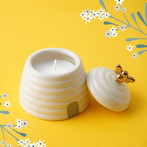Può includere: Portacandele in ceramica bianca a forma di alveare con coperchio. Il coperchio ha un design di ape dorata. La candela è bianca ed è all'interno dell'alveare.