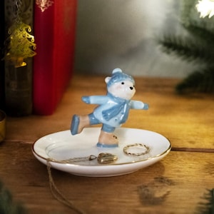 May include: A white ceramic ring dish with a blue and white ceramic bear figurine skating on top. The bear is wearing a blue hat and scarf. There are two gold rings and a gold heart charm on the dish.