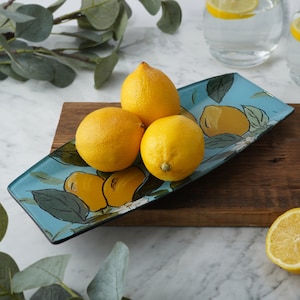 Puede incluir: Una bandeja de servir de vidrio azul con un diseño de limón, con cuatro limones amarillos en la parte superior. La bandeja está sobre una superficie de madera.