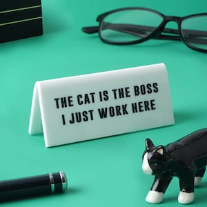 Peut inclure: Un panneau blanc avec du texte noir qui dit "The cat is the boss I just work here". Le panneau est posé sur une surface turquoise avec une figurine de chat noir au premier plan.