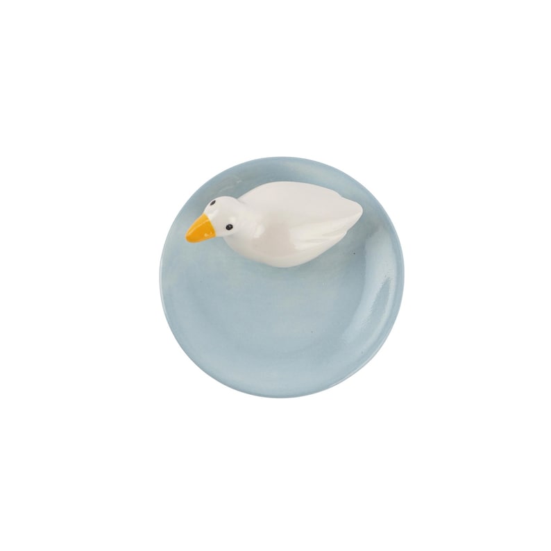 May include: A white ceramic duck with an orange beak floats on a light blue, round, glass-like object. The duck is detailed with black eyes. The object is a decorative item, possibly a paperweight or ornament.