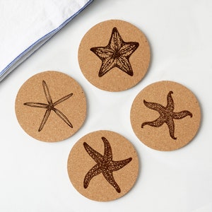 May include: Four round cork coasters with dark brown starfish designs. Each coaster features a different starfish illustration. A white and blue striped cloth is in the upper left corner. The coasters are on a white surface.