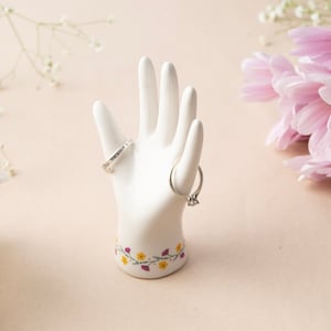 以下が含まれることがあります： 花柄のディテールが施された白いセラミック製のハンドリングホルダー。2つの銀色のリングが指に飾られています。