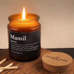 May include: A lit candle in a glass jar with a black label that reads "Mamil (noun)". The label includes a definition and the phrase "The Dapper Chap EST. 2014" is on the cork lid. Matches are next to the candle.