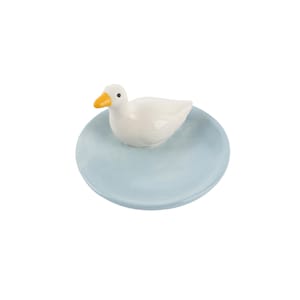 May include: A ceramic duck figurine with a yellow beak and black eyes sits atop a light blue, round dish. The duck is white and the dish has a smooth surface. This decorative item is suitable for home decor.