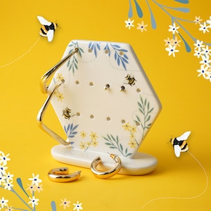 Puede incluir: Un expositor hexagonal de joyería de cerámica blanca con un diseño de abejas y flores. El expositor tiene varios agujeros para pendientes y una base para anillos. Dos aros de oro se muestran en el expositor. Dos abeilles vuelan en el fondo.