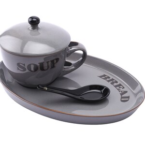May include: A grey ceramic soup bowl with a lid and a matching grey oval tray. The bowl has the word "SOUP" printed on the side. The tray has the word "BREAD" printed on the side. A black spoon is on the tray.