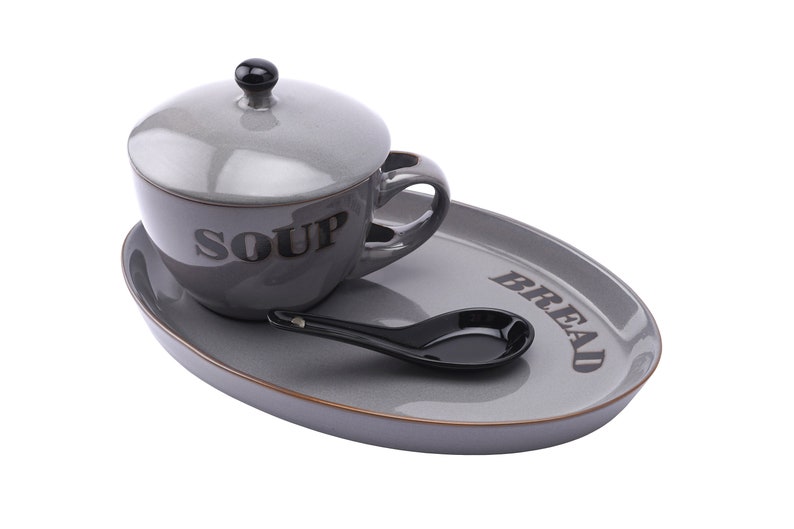 May include: A grey ceramic soup bowl with a lid and a matching grey oval tray. The bowl has the word "SOUP" printed on the side. The tray has the word "BREAD" printed on the side. A black spoon is on the tray.