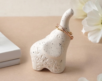 May include: White ceramic elephant ring holder with black speckles and floral details. Two gold rings are on the elephant's trunk.