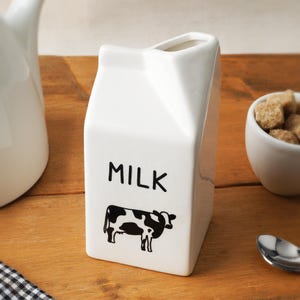 Może przedstawiać: Biały ceramiczny wazon w kształcie kartonu po mleku ze słowem "MILK" w czarnych literach i czarno-białą ilustracją krowy. Wazon stoi na drewnianej powierzchni.