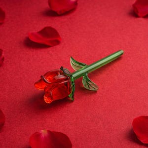 Puede incluir: Una rosa de cristal con una flor roja y un tallo y hojas verdes. La rosa está sobre un fondo rojo con pétalos de rosa rojos esparcidos, creando una exhibición romántica. Objeto decorativo.