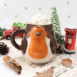 Könnte beinhalten: Eine braune Keramiktasse in Form eines Rotkehlchens mit weißem Rand und weißem Henkel. Die Tasse ist gefüllt mit einem weißen, cremigen Getränk, das mit Marshmallows bedeckt ist. Die Tasse steht auf einem Notenblatt mit einem Kiefernzweig, einem roten und weißen Briefkasten und Zimtstangen im Hintergrund.