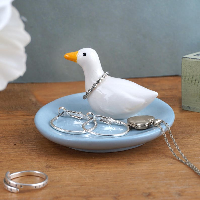 May include: A white ceramic duck figurine with an orange beak and a silver bracelet around its neck sits on a light blue dish. Silver hoop earrings, a heart-shaped locket, and a ring are also on the dish.