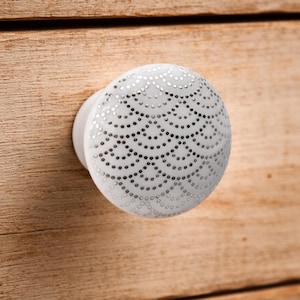 May include: A round white ceramic cabinet knob featuring a repeating pattern of small silver dots arranged in overlapping scalloped arches. The knob has a smooth, glossy finish and a simple, functional shape designed for furniture drawers or doors.