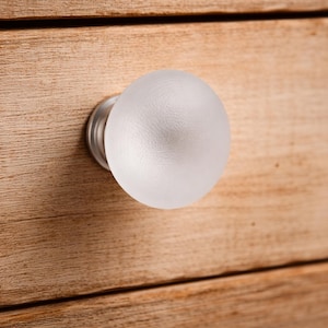 May include: A round, frosted glass cabinet knob with a smooth, spherical surface and a silver-toned metal base. The knob is attached to a light-colored wooden drawer front with a visible grain pattern, showcasing a simple and functional design.