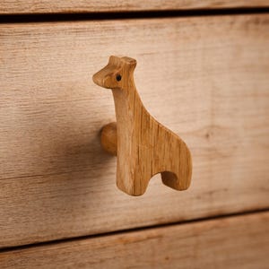 Op de afbeelding: Een lichtbruine houten ladeknop in de vorm van een giraf. De knop heeft een natuurlijke houtnerftextuur en een kleine, donkere, cirkelvormige stip die het oog voorstelt. Het is gemonteerd op een lichtgekleurd houten ladefront met horizontale nerf.