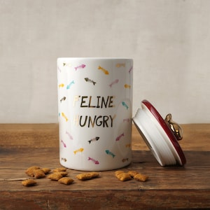 Op de afbeelding: Witte keramische kattenvoerpot met een rood deksel en een gouden kattenpootknop. De pot heeft een gouden inscriptie met de tekst "Feline Hungry" en is versierd met een patroon van kleurrijke visgraten.