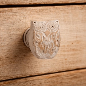 Op de afbeelding: Een houten, gebeeldhouwde uilvormige trekker met een verweerde witte afwerking. De ogen en veren van de uil zijn gedetailleerd met gebeeldhouwde lijnen. De trekker is gemonteerd op een lichtbruine houten lade.