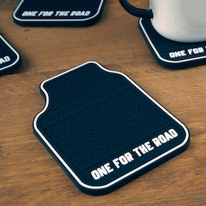 May include: Black, car-mat-shaped coasters with white trim and the text "ONE FOR THE ROAD." The coasters have a textured surface and are on a wooden table. A white mug sits on one of the coasters.
