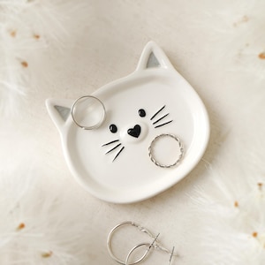 May include: White ceramic cat-shaped trinket dish with black painted facial features. The dish has two silver rings inside.