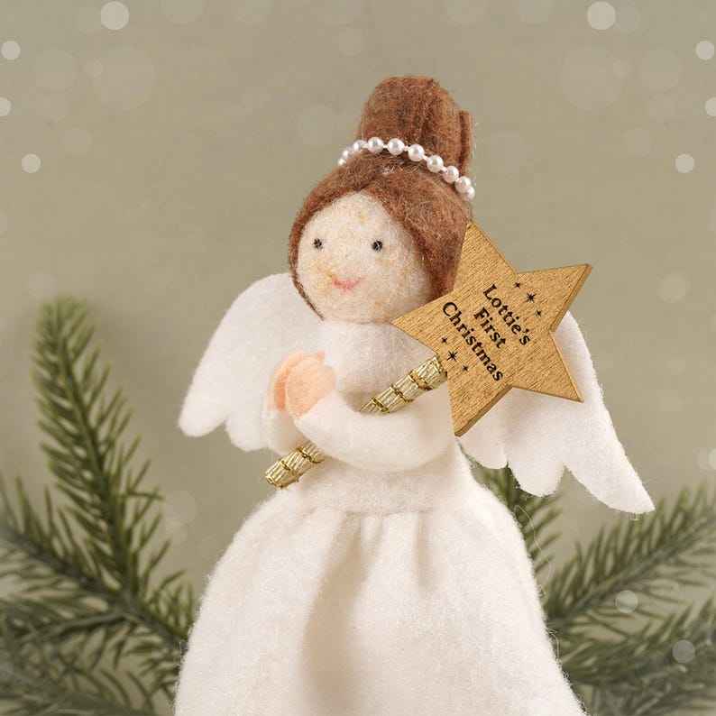 May include: A felt angel Christmas ornament with brown hair, a pearl halo, and white wings. The angel holds a wooden star wand with the text "Lottie's First Christmas". The angel wears a white dress and has a light skin tone.