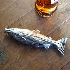Könnte beinhalten: Ein verchromter Flaschenöffner in Fischform. Der Öffner hat ein detailliertes Schuppenmuster und Flossendesign. Der Flaschenöffner liegt auf einer Holzoberfläche, mit einer Flasche und einem Glas im Hintergrund.