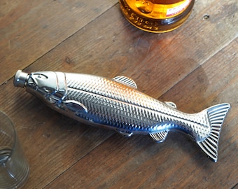 May include: A chrome-finished fish-shaped bottle opener. The opener has a detailed scale pattern and fin design. The bottle opener is lying on a wooden surface, with a bottle and a glass visible in the background.