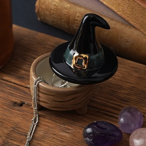 May include: A black witch hat shaped trinket box with a green and gold band. The box is open and a silver chain necklace is inside. The box is sitting on a wooden surface with three amethyst stones.